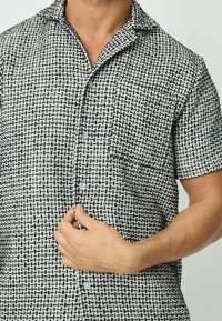 Chemise à manches courtes avec un motif tissé noir et blanc. Présente un col, des boutons et une poche sur la poitrine. Le tissu a une apparence texturée.