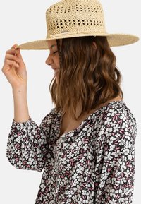 Slomovaný sluneční klobouk s širokým krempou a otevřeným vzorem. Modelka má na sobě květinový top v černé, bílé a růžové. Klobouk drží jednou rukou.