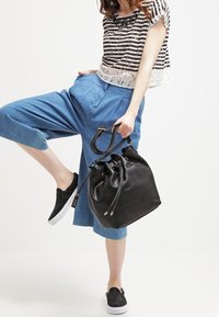 Sort perforeret læderbucket taske med snørrelukning og dobbelte hanke, parret med blå culottes og en stribet top.