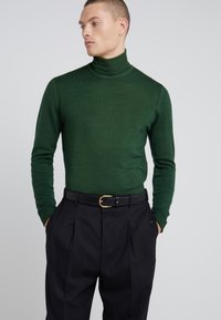 Suéter verde de cuello alto hecho de tela suave, diseño ajustado, mangas largas, combinado con pantalones negros de cintura alta y un cinturón negro con herrajes dorados.