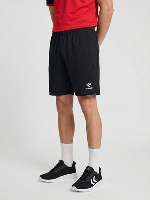 Mand iført sorte Hummel-shorts, sorte sneakers med hvide såler, hvide sokker og en rød t-shirt med korte ærmer, står på en ensfarvet baggrund.