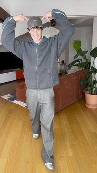 Grå fleecejakke med lynlås, sort kasket, grå teksturerede bukser og sølvfarvede sneakers. Indendørs omgivelser med trægulv og grøn plante i baggrunden.