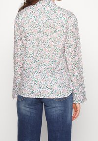 Blouse à manches longues en tissu léger, présentant un motif floral coloré sur fond blanc. Col rond et ourlet arrondi.