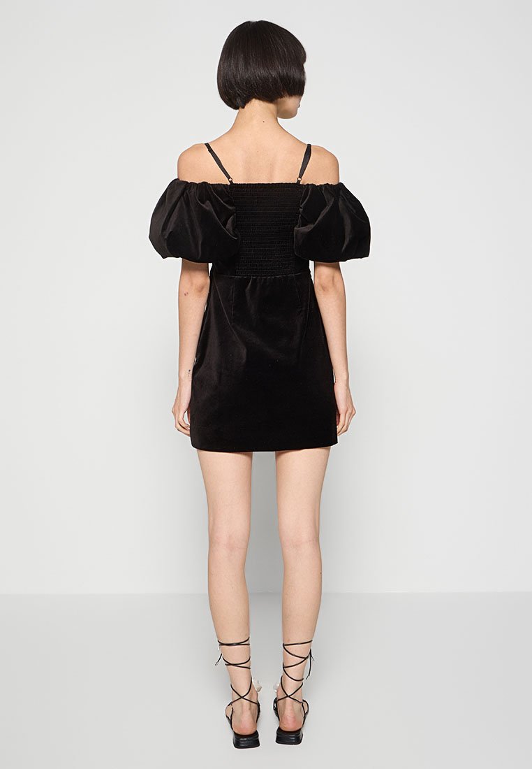Robe mini en velours noir avec des manches bouffantes et un corsage smocké. Présente une silhouette ajustée et une longueur au-dessus du genou. Talons noirs à lanières.
