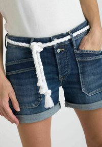 Shorts en denim avec un délavage bleu clair, ourlets roulés, et une ceinture en corde tressée blanche avec des pompons, présentés avec une main sur la poche.