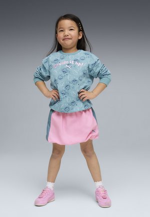 Młoda dziewczyna stoi pewnie z rękami na biodrach, ubrana w niebieską bluzę z napisem „Śnij to!” oraz różową spódnicę i buty.