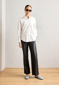 Camicia bianca a bottoni, pantaloni larghi in pelle nera e ballerine blu a fantasia. Il modello indossa occhiali da sole neri. Ambientazione minimalista con pavimento in legno.