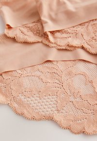 Beżowa koronka z kwiatowymi wzorami i falistymi krawędziami nałożona na gładki, dopasowany beżowy materiał, ukazująca misterną fakturę.