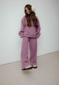 Fioletowy dzianinowy sweter i pasujące spodnie, o luźnym kroju i miękkiej fakturze. Białe nauszniki z sztucznego futra dopełniają wygląd.