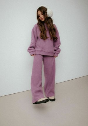 Fioletowy dzianinowy sweter i pasujące spodnie, o luźnym kroju i miękkiej fakturze. Białe nauszniki z sztucznego futra dopełniają wygląd.