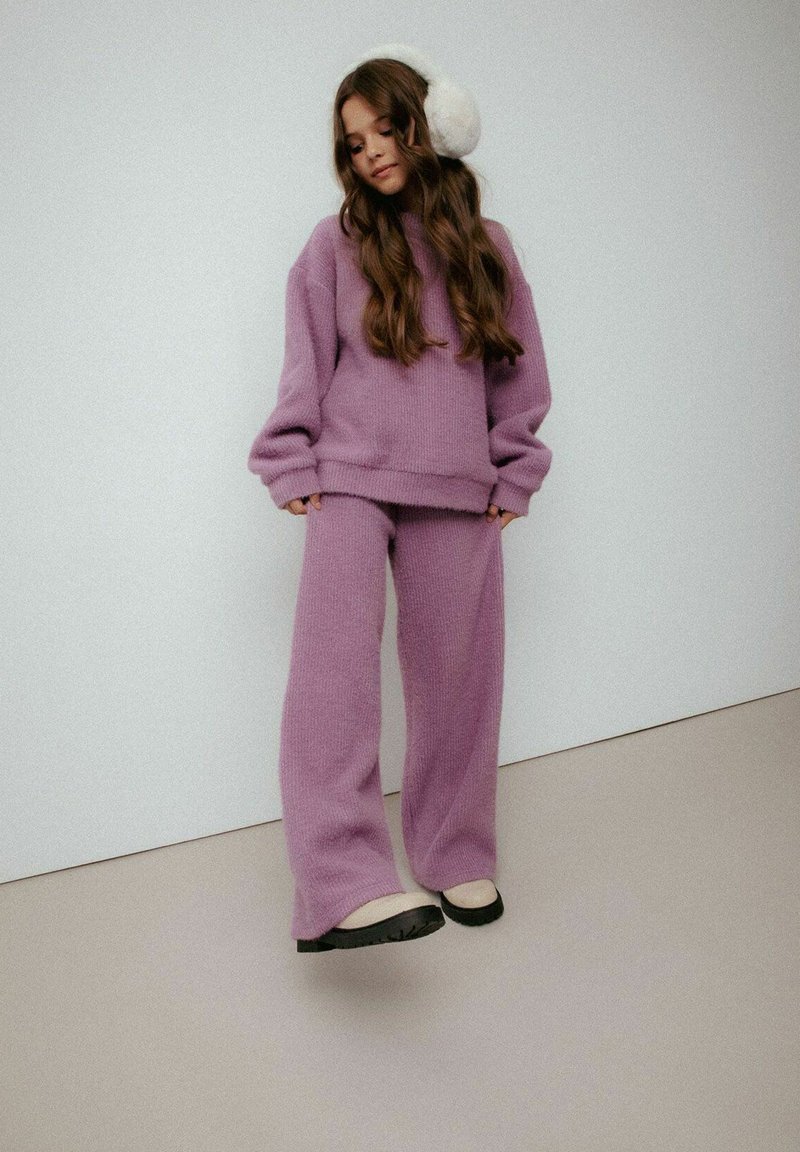 Fioletowy dzianinowy sweter i pasujące spodnie, o luźnym kroju i miękkiej fakturze. Białe nauszniki z sztucznego futra dopełniają wygląd.
