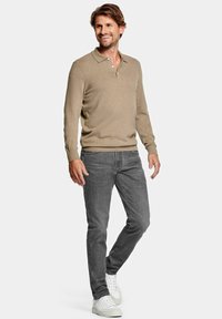 Beige poloshirt met lange mouwen en twee knopen, gecombineerd met grijze jeans en witte sneakers. De outfit heeft een casual en aangesloten uitstraling.
