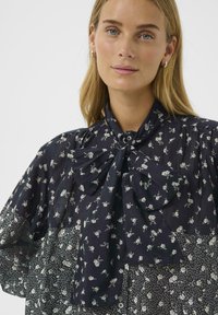 Blonde woman with blue eyes wearing silver hoop earrings and a navy blouse with white floral pattern and large bow tied at collar.