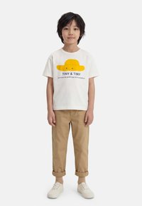 Παιδί με μαύρα μαλλιά που φοράει λευκό μπλουζάκι με κίτρινο σχέδιο καπέλου και κείμενο, μπεζ παντελόνι γυρισμένο, μπεζ κάλτσες και λευκά αθλητικά παπούτσια να στέκεται.