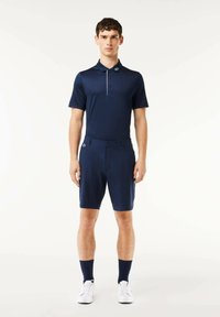 Polo de color marino con mangas cortas, que presenta un cuello y una tapeta de botones. Combinado con pantalones cortos a juego, zapatillas blancas y calcetines de caña corta en color marino.