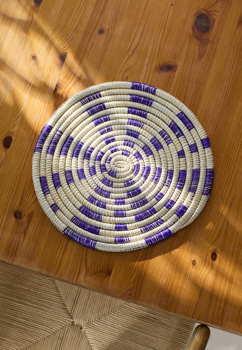 Round braided placemat made of natural fibers, featuring alternating purple and white stripes, on a wooden table with a textured surface.