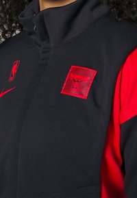 Veste de sport noire et rouge avec un patch du logo des Chicago Bulls et les logos NBA et Nike sur la poitrine, portée par une personne aux cheveux bouclés.