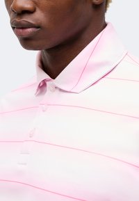 Κοντινό πλάνο ενός ατόμου που φοράει ένα απαλό ροζ πόλο μπλουζάκι με λεπτές οριζόντιες ρίγες και τρία κουμπιά κοντά στον γιακά.