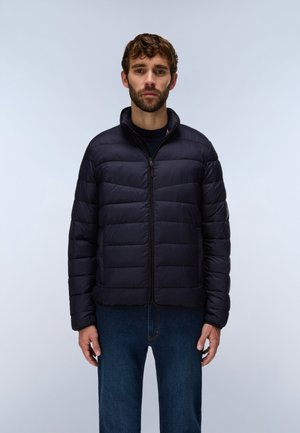 Homme aux cheveux courts et bouclés portant une veste noire matelassée zippée et un jean bleu, debout devant un fond clair uni.