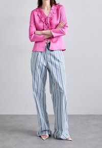 Blusa rosa a volani con maniche lunghe abbinata a pantaloni a gamba larga blu e bianchi a righe. Il modello si trova su una superficie grigia.