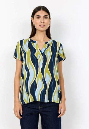 Vrouw draagt een blouse met korte mouwen met blauwe, groene en marineblauwe golvende strepen, gecombineerd met donkerblauwe spijkerbroek tegen een witte achtergrond.