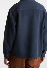 Henkilö, jolla on yllään tummansininen teksturoitu pitkähihainen paita ja ruskeat housut, näkyy takaa kädet rentoina sivuilla.