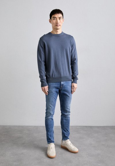 Pull gris à col rond en tissu tricoté doux, associé à un jean bleu clair et des baskets beiges avec des semelles en caoutchouc. Design simple, sans motifs.
