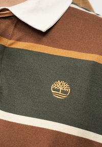 Κοντινό πλάνο σε πουκάμισο με γιακά με καφέ, πράσινες και κίτρινες ρίγες και κεντημένο λογότυπο δέντρου στο στήθος.