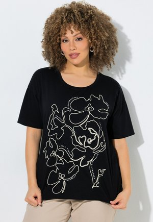 Kvinde med krøllet hår iført en sort T-shirt med hvid abstrakt blomsterlinjekunst, stående mod en hvid baggrund.