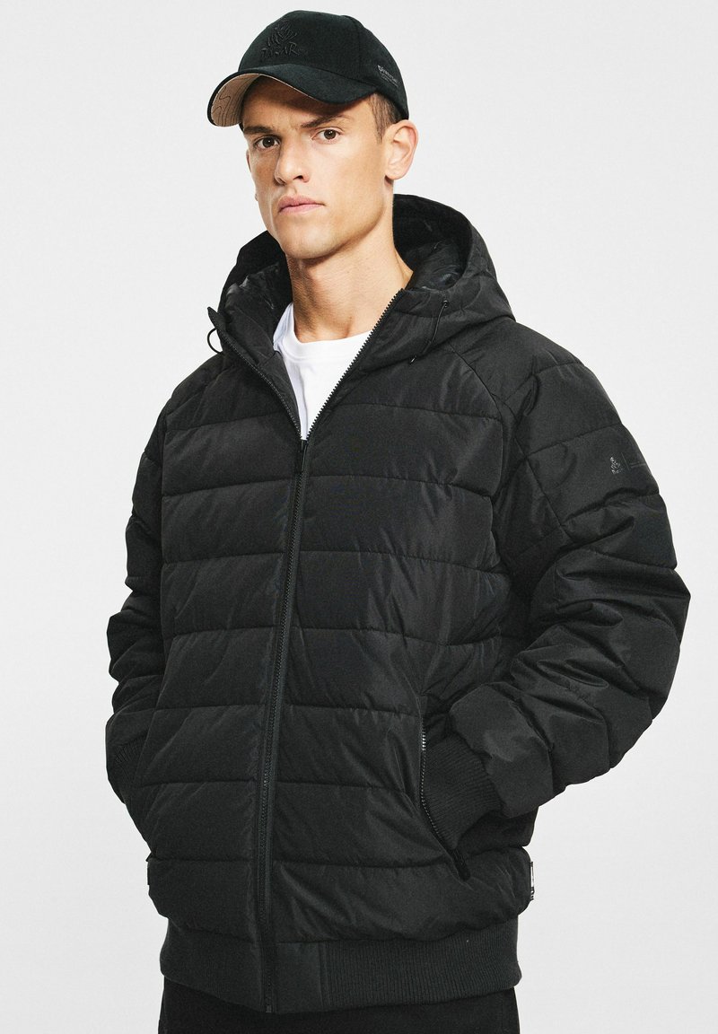 Czarna puchowa kurtka z kapturem, z pikowanym wzorem, zamkiem błyskawicznym z przodu, elastycznymi ściągaczami na rękawach i bocznymi kieszeniami. Noszona na białej koszuli.