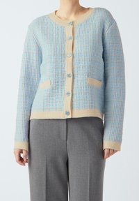 Sweter zapinany na guziki w jasnoniebieskim kolorze z kremowymi akcentami. Posiada okrągły dekolt, zapięcie na guziki oraz dwie przednie kieszenie. Miękka dzianinowa tkanina.