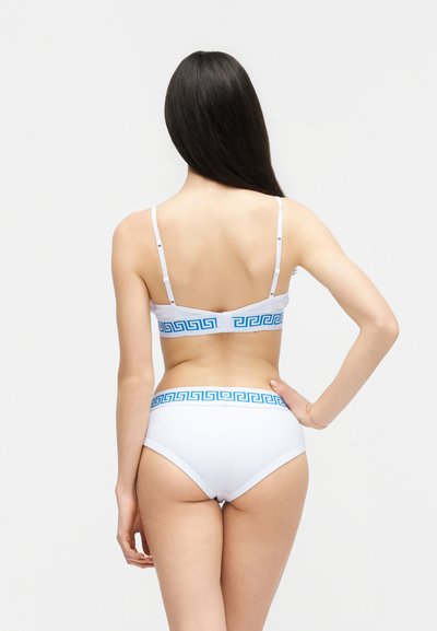 Γυναίκα που φαίνεται από πίσω, φοράει λευκό σουτιέν και σλιπ με μπλε διακοσμητικό μοτίβο ελληνικού κλειδιού στις ταινίες, στέκεται μπροστά σε απλό φωτεινό φόντο.