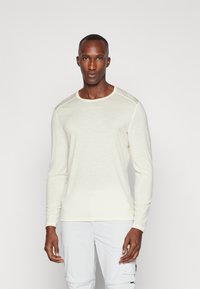 Chemise en tricot crème clair à manches longues avec un col rond et des coutures d'accent au niveau des épaules, associée à un pantalon blanc. Texture lisse.