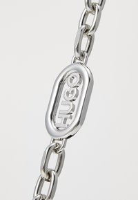 Zilveren metalen kettingarmband met een ovale schakel met uitgesneden "HUGO" tekst, een gladde textuur en een gepolijste afwerking.