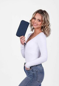 Denimová peněženka tmavě námořnické barvy, obdélníkového tvaru, hladké struktury a se zipovým uzavřením, držená proti modelu ve bílé blůze.