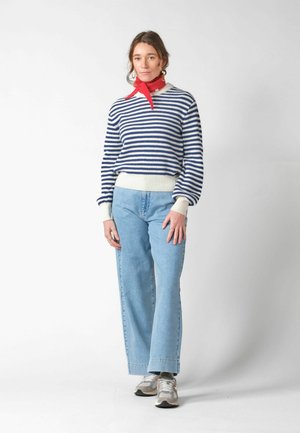 Kvinde stående iført en blå og hvidstribet sweater, lyseblå jeans, grå sneakers og et rødt halstørklæde foran en ensfarvet baggrund.