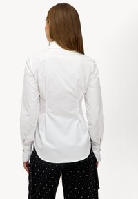 Camisa blanca de manga larga con diseño entallado, pliegues en la parte posterior para dar forma y dobladillo curvo. Combinada con pantalones negros estampados.