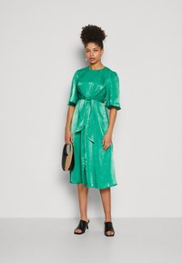 Türkisfarbenes Satin-Kleid mit kurzen Puffärmeln, tailliert, knielang, kombiniert mit schwarzen offenen Mules und einer zweifarbigen Handtasche.
