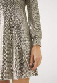 Robe en sequins argentés avec manches longues, de forme évasée et surface texturée reflétant la lumière. Présente un poignet ajusté et un ourlet lisse.