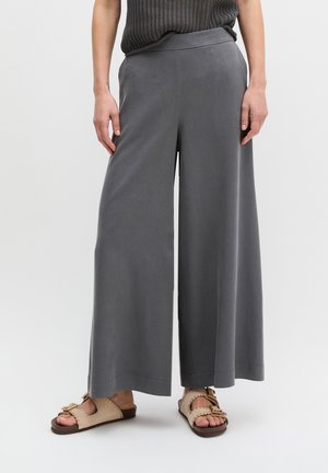 Osoba ubrana w luźne, szare spodnie o szerokich nogawkach, beżowe sandały z klamrami, stojąca na jednolitym jasnym tle.