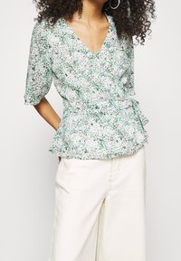 Blusa avvolgente floreale in verde e bianco con scollo a V, maniche corte, orlo peplum e una cravatta coordinata in vita. Tessuto leggero.