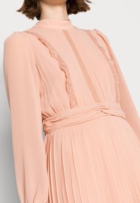 Robe en chiffon pêche avec des manches longues, des accents en dentelle le long de l'encolure et une jupe plissée. Présente une taille cintrée avec une ceinture nouée.