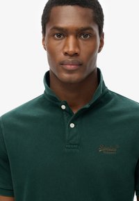 Polo shirt verde scuro con finitura testurizzata, dotato di un colletto classico, tre bottoni e un piccolo logo sul lato sinistro del petto.