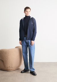 Felpa zip-up blu navy con fodera grigia, indossata sopra un dolcevita nero, abbinata a jeans leggermente svasati blu chiaro e scarpe nere. Sedia beige testurizzata nelle vicinanze.