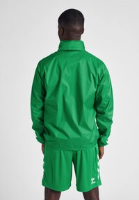 Grön vindjacka med hög krage, långa ärmar och slät textur; matchad med gröna shorts med vita sidodetaljer.