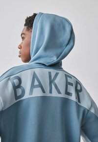 Hellblauer Kapuzenpullover mit einem kontrastierenden, dunkler blauen Einsatz. Großes "BAKER"-Schriftzug auf dem oberen Rücken. Weiche Textur.