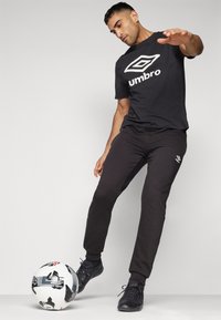 Čierne bavlnené tričko s bielym logom Umbro, spárované s čiernymi teplákmi. Model je postavený s futbalovou loptou pri nohách.