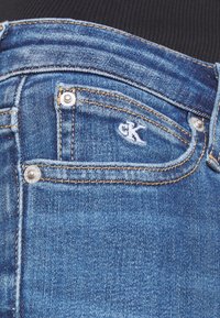 Modré džíny z denimu s přední kapsou a stříbrným patentem, na kterém je vyšitá logo CK. Ošuntělá textura, slim fit design.