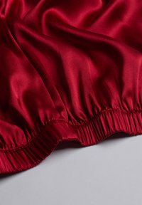 Rode satijnen stof met een gladde textuur, voorzien van een gerimpelde elastische tailleband langs de onderrand. Lichtreflecties geven diepte.