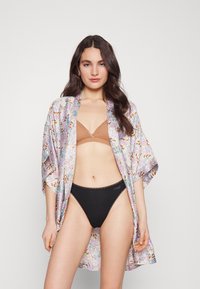 Blommönstrad kimono i lätt tyg, med vida ärmar och pastellfärger, bärs över en brun triangulär bikiniöverdel och svarta underkläder.